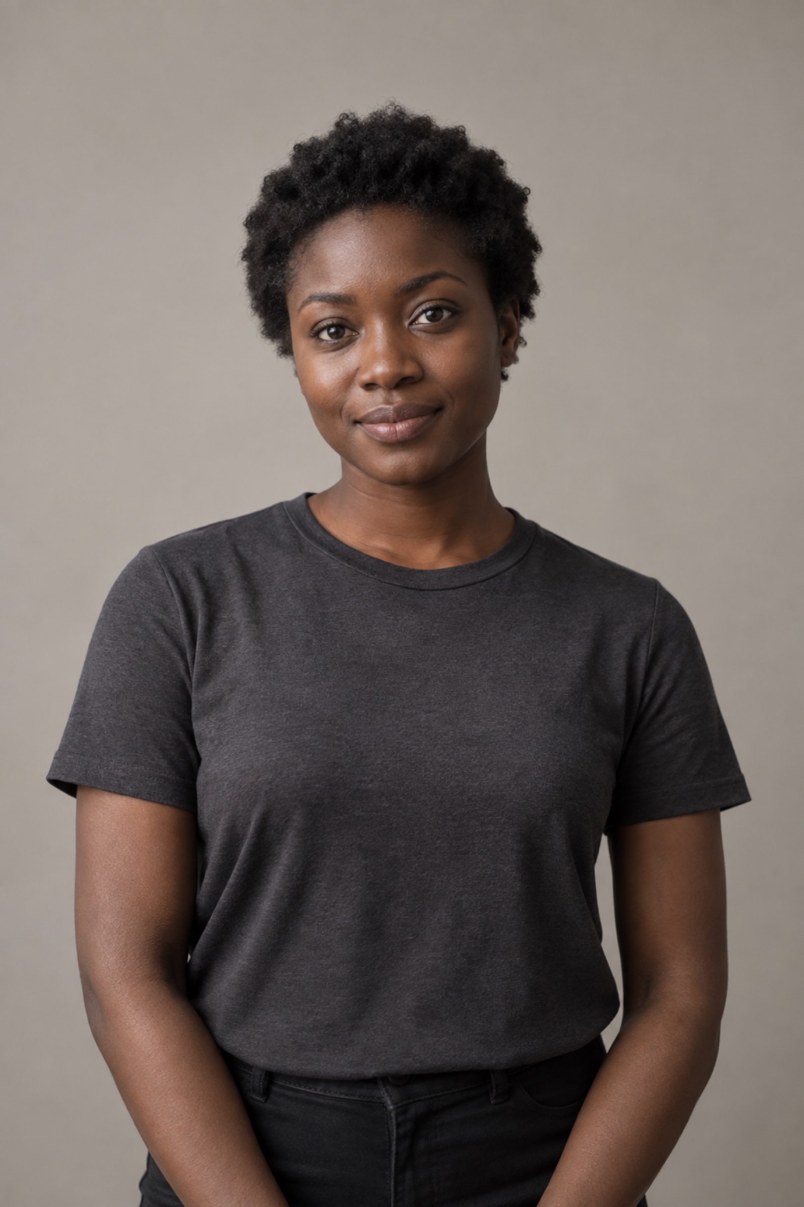 Studio portrait of a woman in their 20s with short natural coily hair, wearing a charcoal shirt, against a warm grey background.