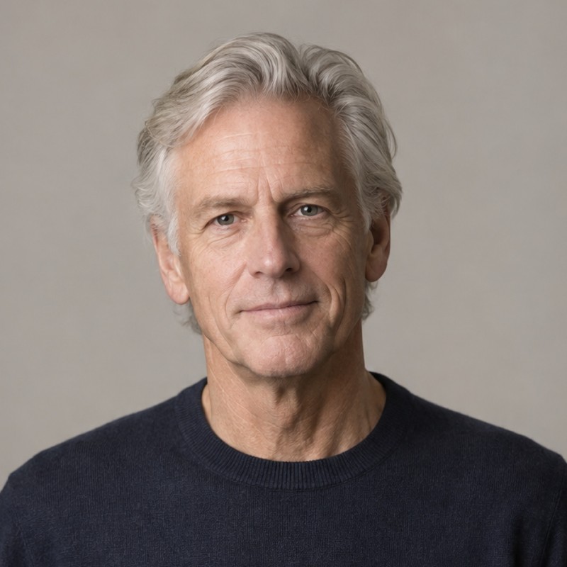 Head and shoulders of a man in their 60s with medium-length swept-back silver hair, wearing a navy sweater, against a warm grey background.