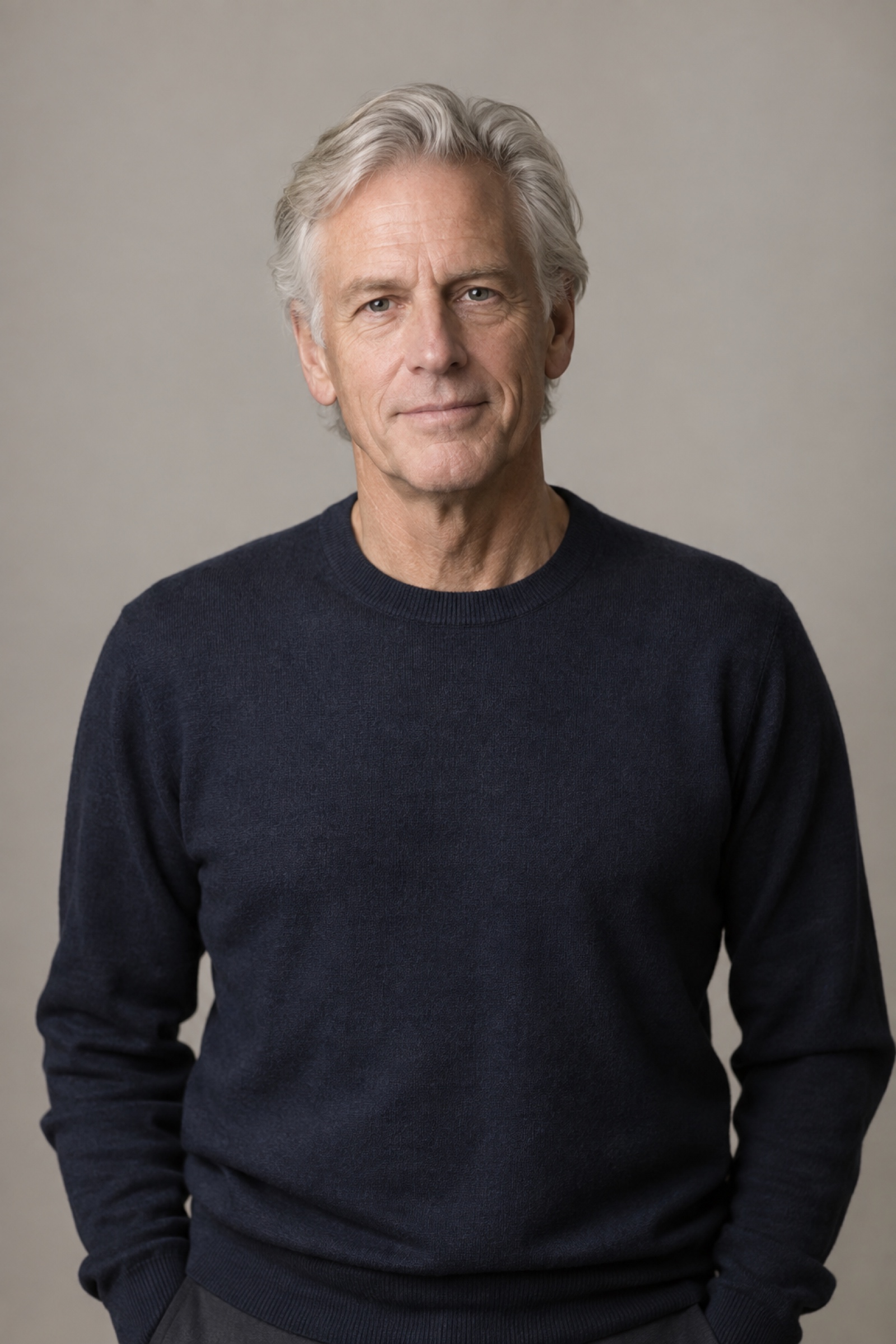 Studio portrait of a man in their 60s with medium-length swept-back silver hair, wearing a navy sweater, against a warm grey background.