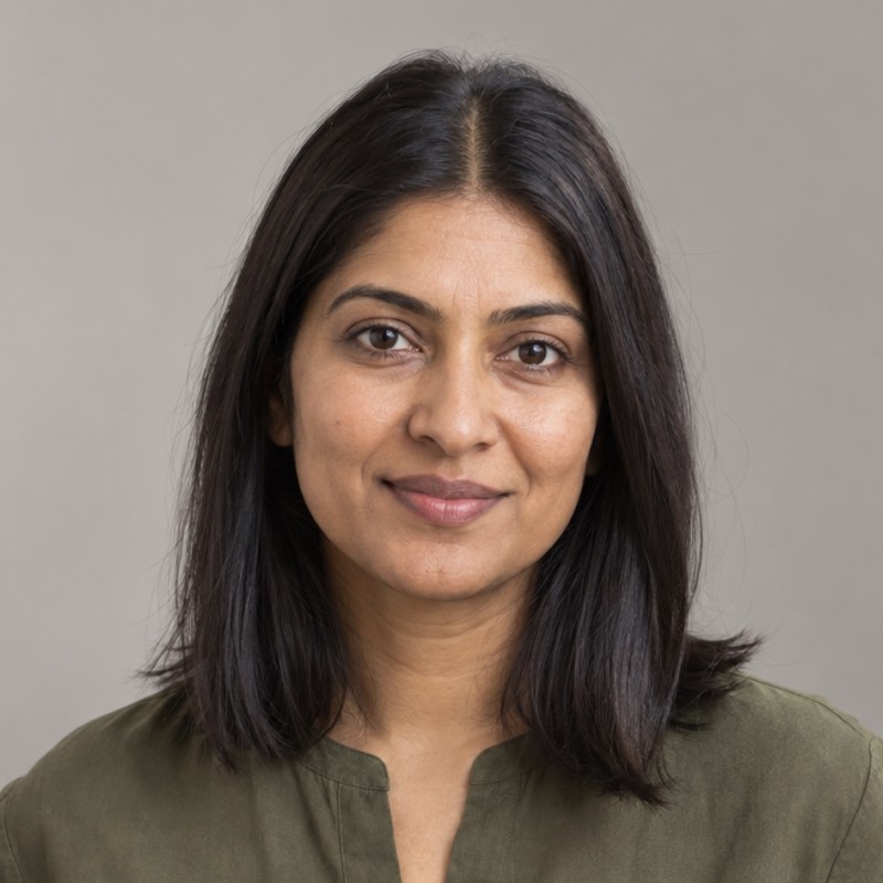 Head and shoulders of a woman in their 30s with straight dark shoulder-length hair with centre part, wearing an olive blouse, against a warm grey background.