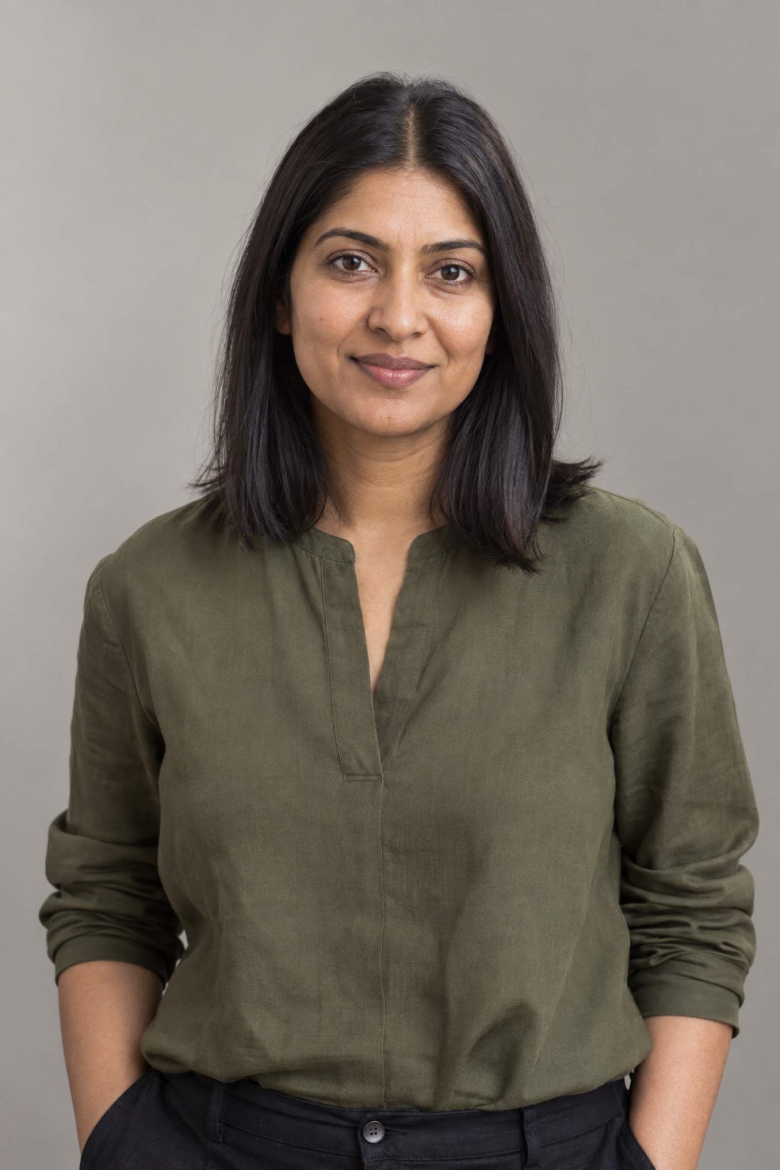 Studio portrait of a woman in their 30s with straight dark shoulder-length hair with centre part, wearing an olive blouse, against a warm grey background.