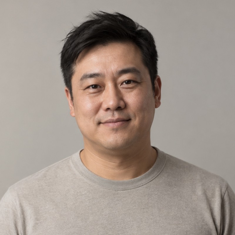 Head and shoulders of a man in their 40s with short dark hair, wearing a stone shirt, against a warm grey background.