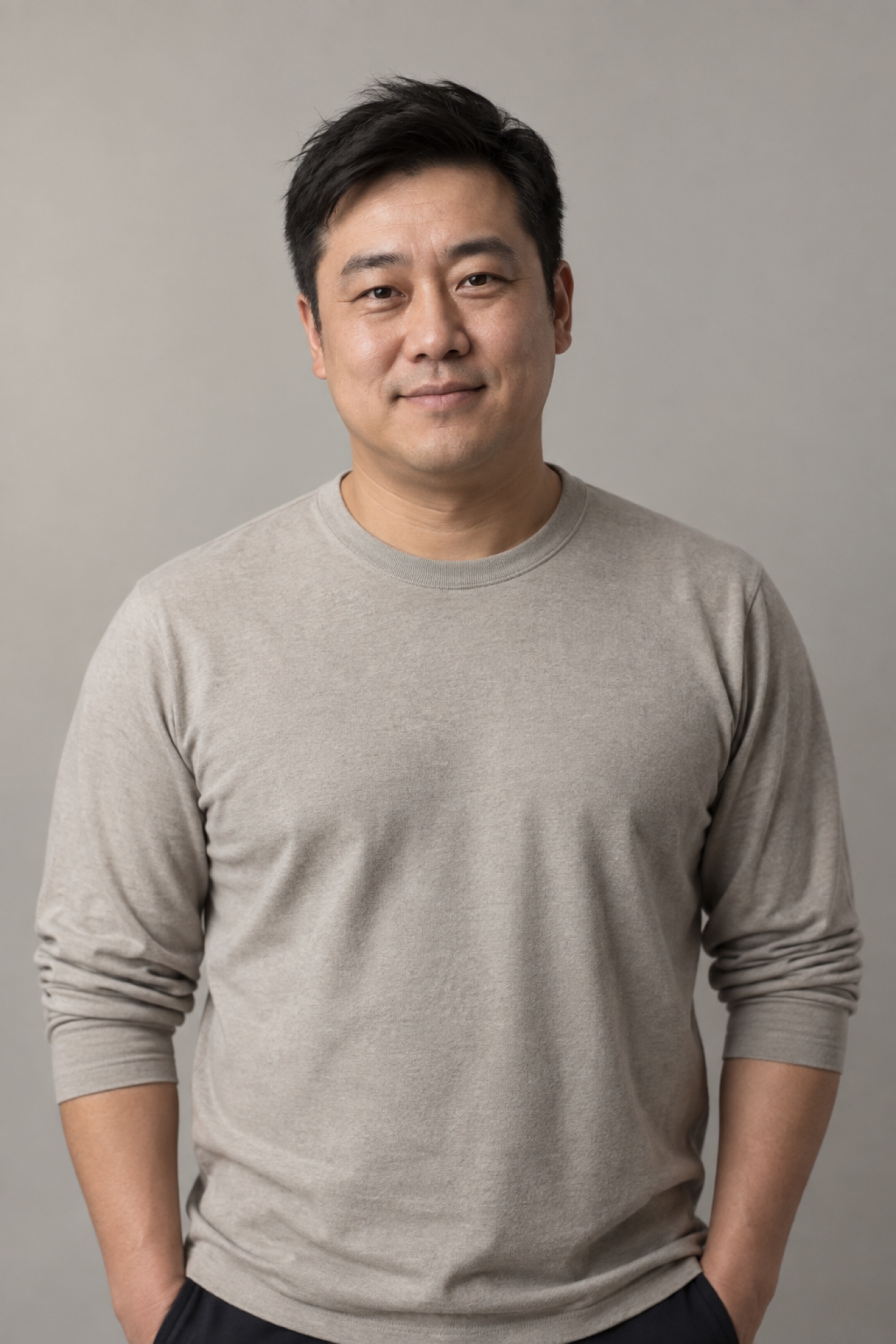 Studio portrait of a man in their 40s with short dark hair, wearing a stone shirt, against a warm grey background.