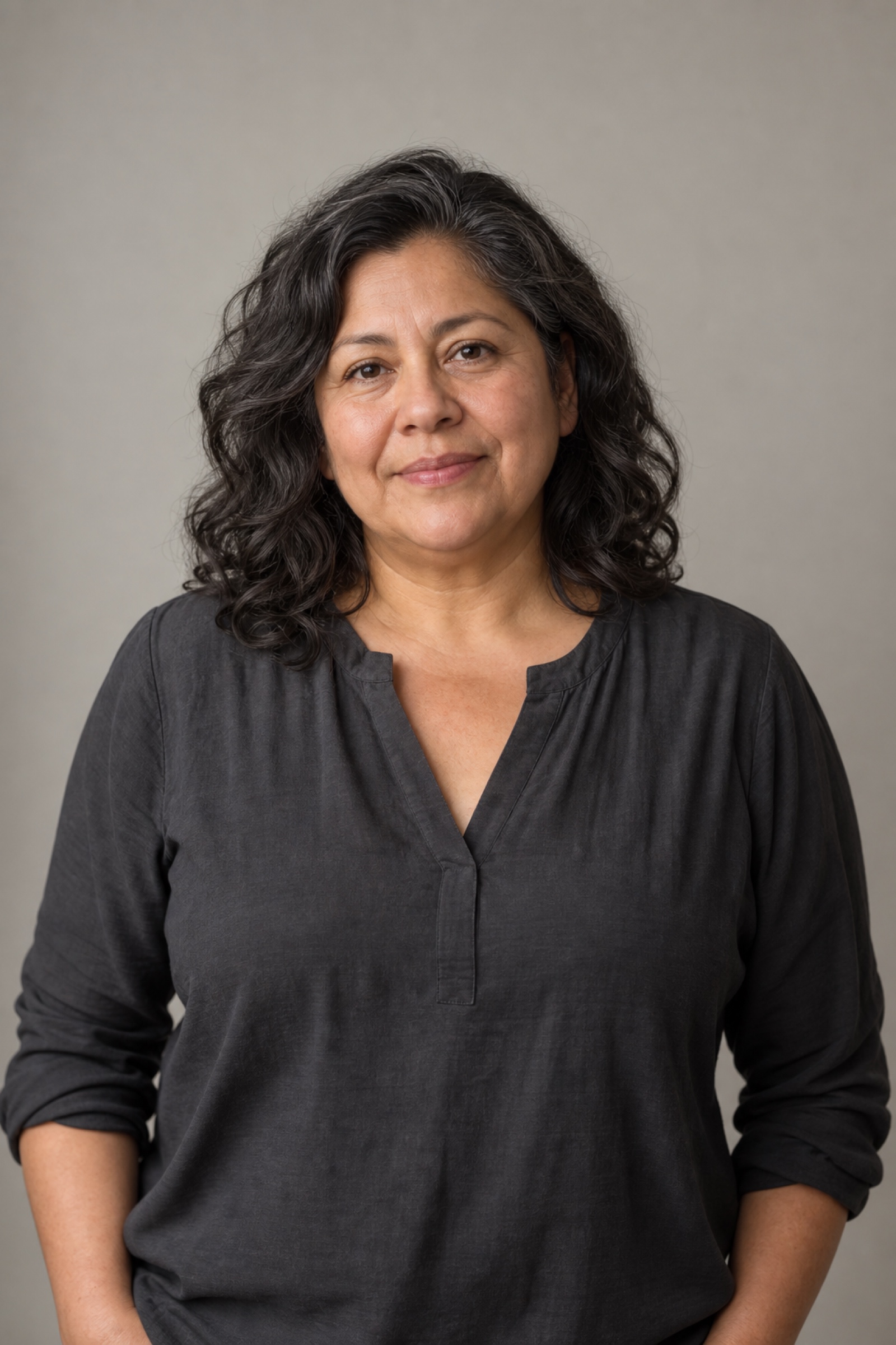 Studio portrait of a woman in their 50s with wavy dark hair to the shoulders with a few grey strands, wearing a charcoal blouse, against a warm grey background.
