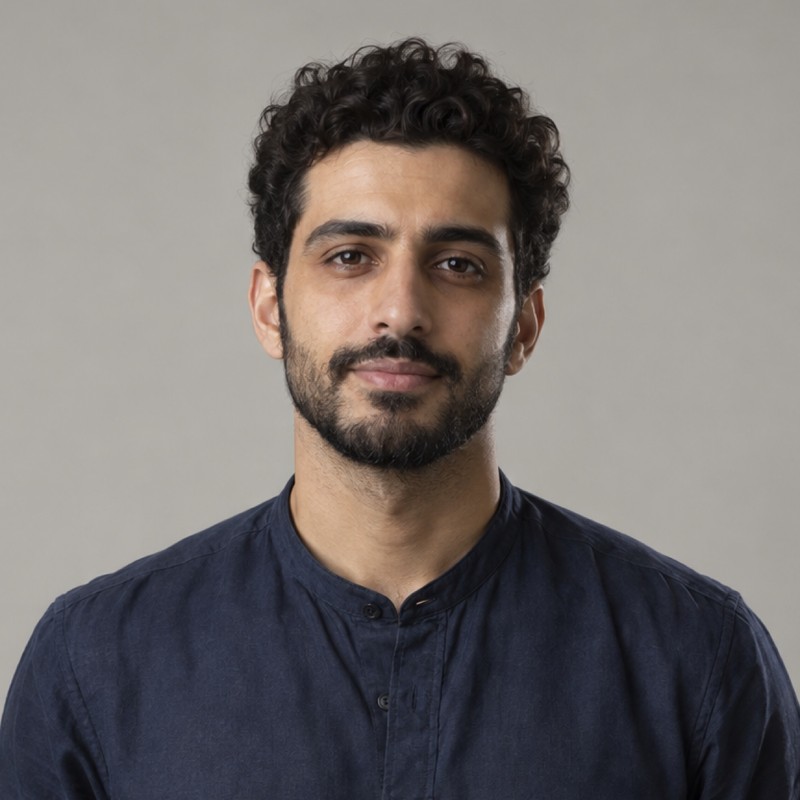 Head and shoulders of a man in their 20s with short curly dark hair and neat short beard, wearing a navy shirt, against a warm grey background.