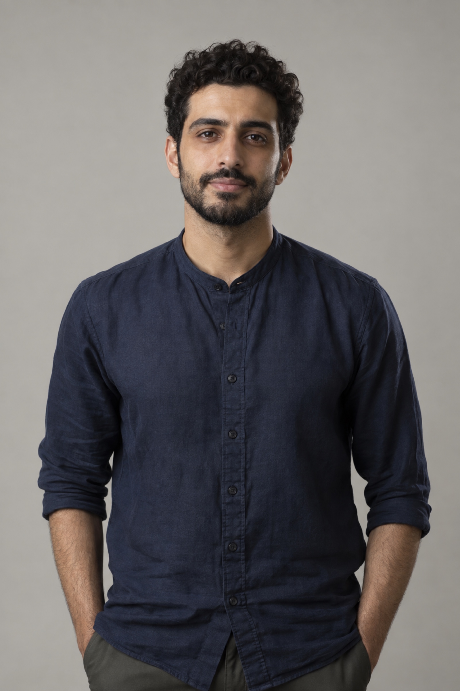 Studio portrait of a man in their 20s with short curly dark hair and neat short beard, wearing a navy shirt, against a warm grey background.