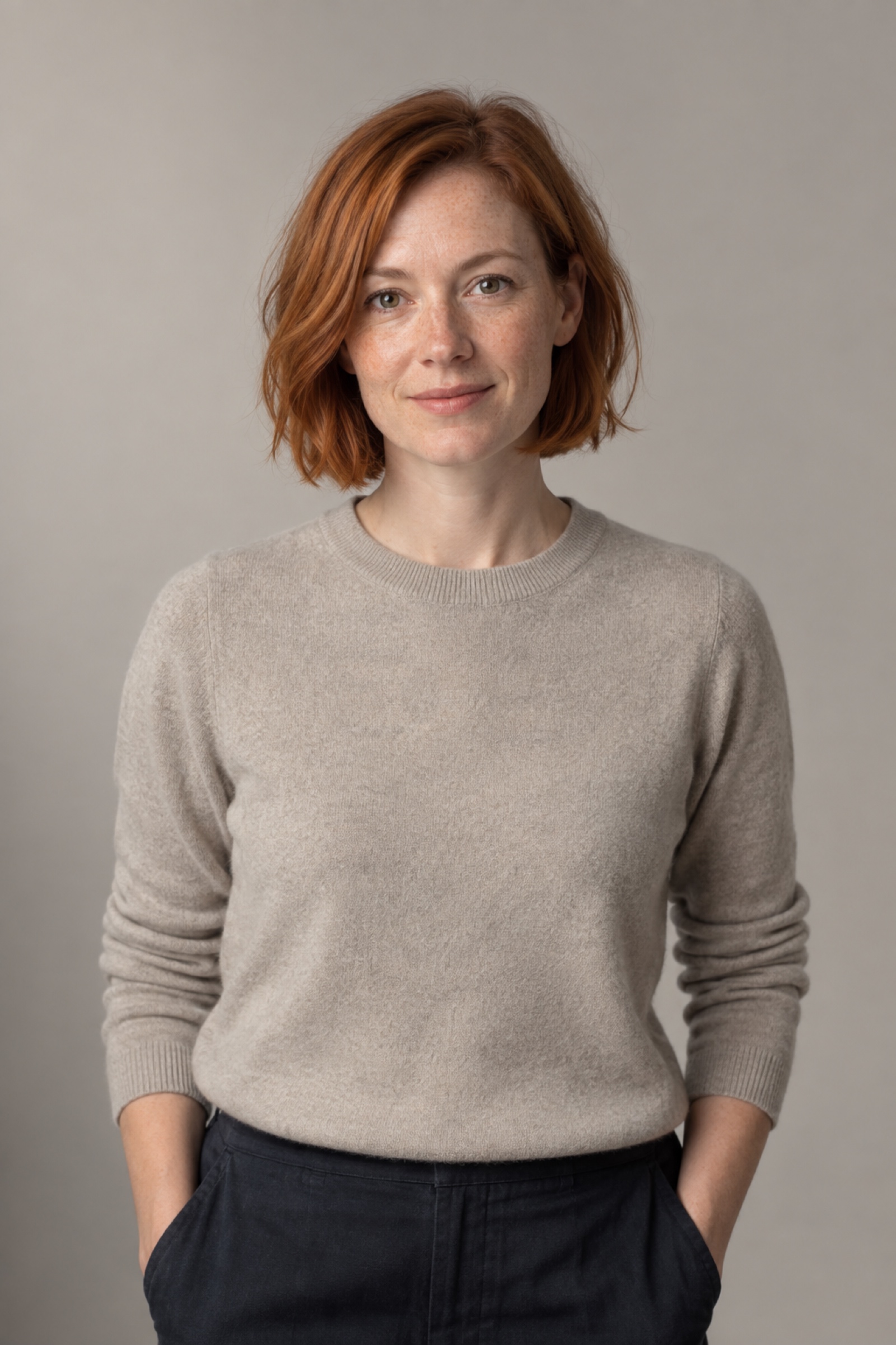Studio portrait of a woman in their 30s with chin-length copper-red bob, wearing a stone sweater, against a warm grey background.