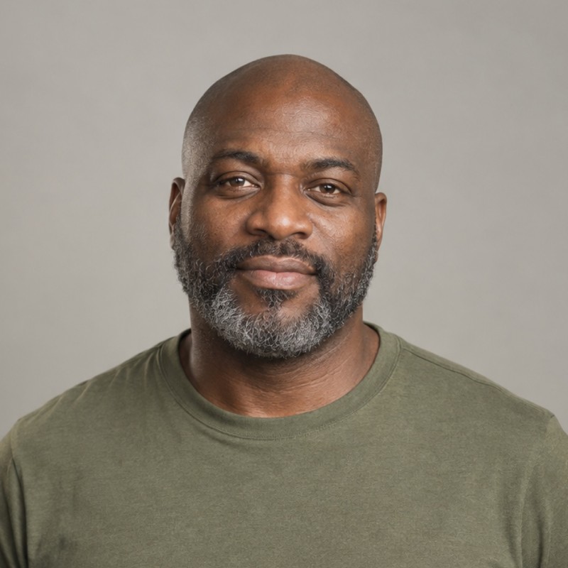 Head and shoulders of a man in their 40s with shaved head, wearing an olive shirt, against a warm grey background.
