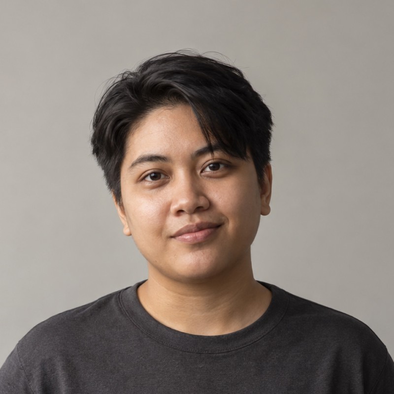 Head and shoulders of a person in their 20s with short straight dark hair with a side part, wearing a charcoal shirt, against a warm grey background.