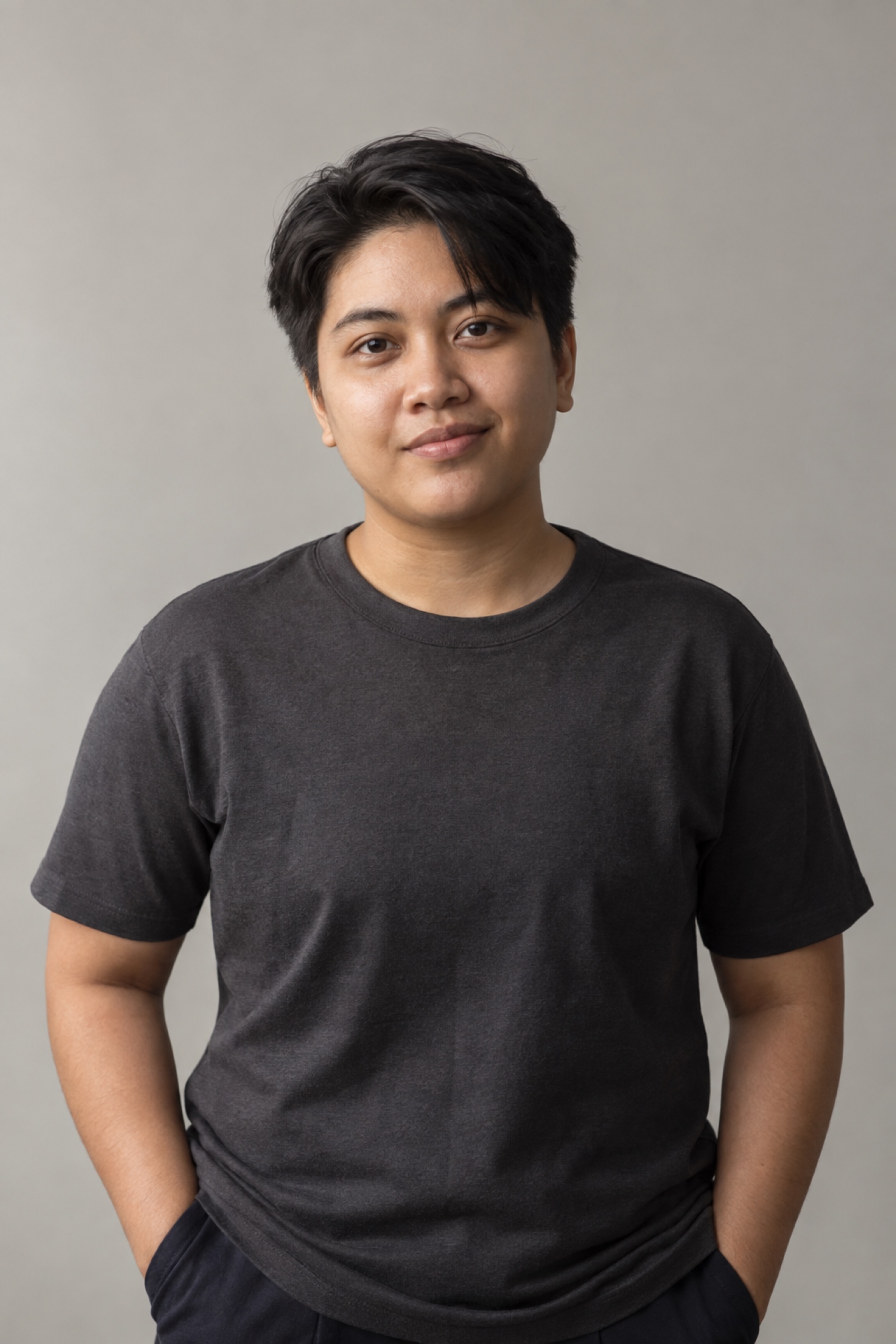Studio portrait of a person in their 20s with short straight dark hair with a side part, wearing a charcoal shirt, against a warm grey background.