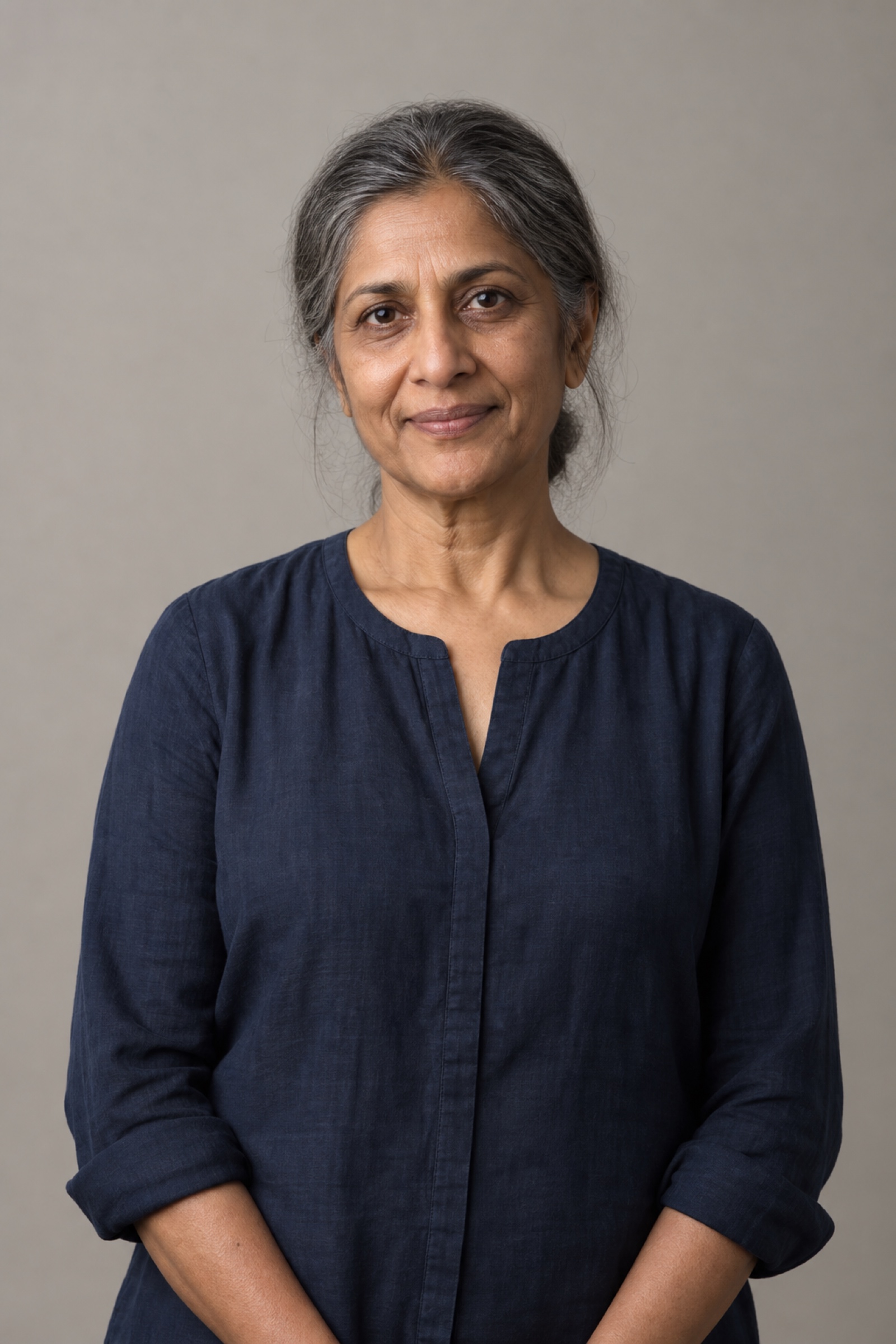 Studio portrait of a woman in their 50s with silver-streaked dark hair loosely tied back, wearing a navy blouse, against a warm grey background.