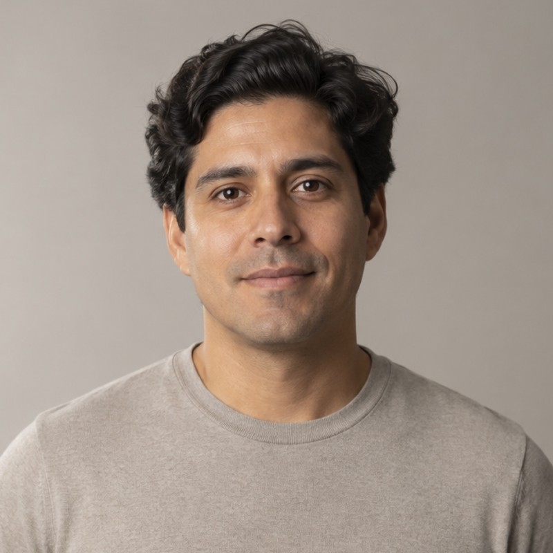 Head and shoulders of a man in their 30s with thick wavy dark hair, wearing a stone shirt, against a warm grey background.