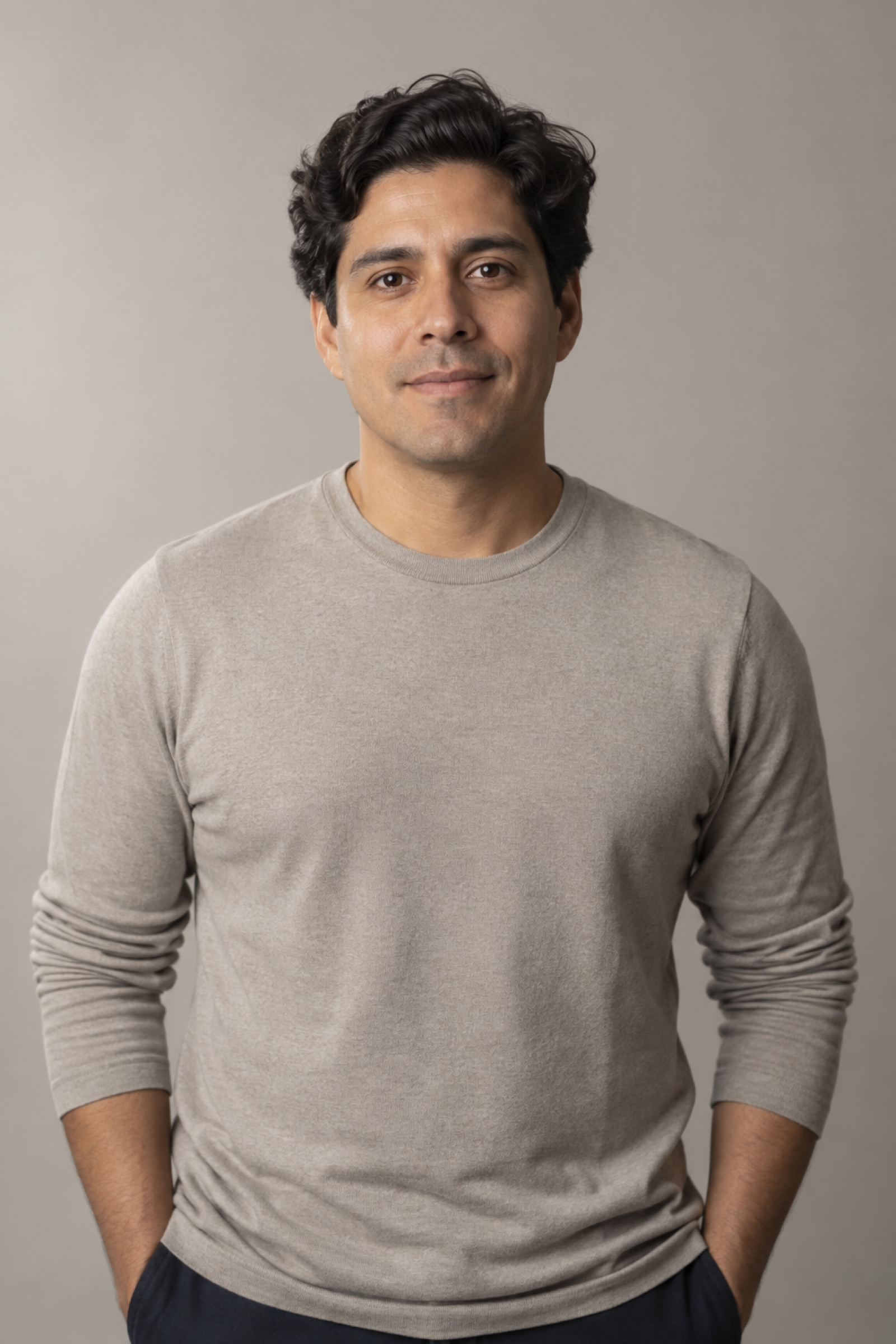 Studio portrait of a man in their 30s with thick wavy dark hair, wearing a stone shirt, against a warm grey background.
