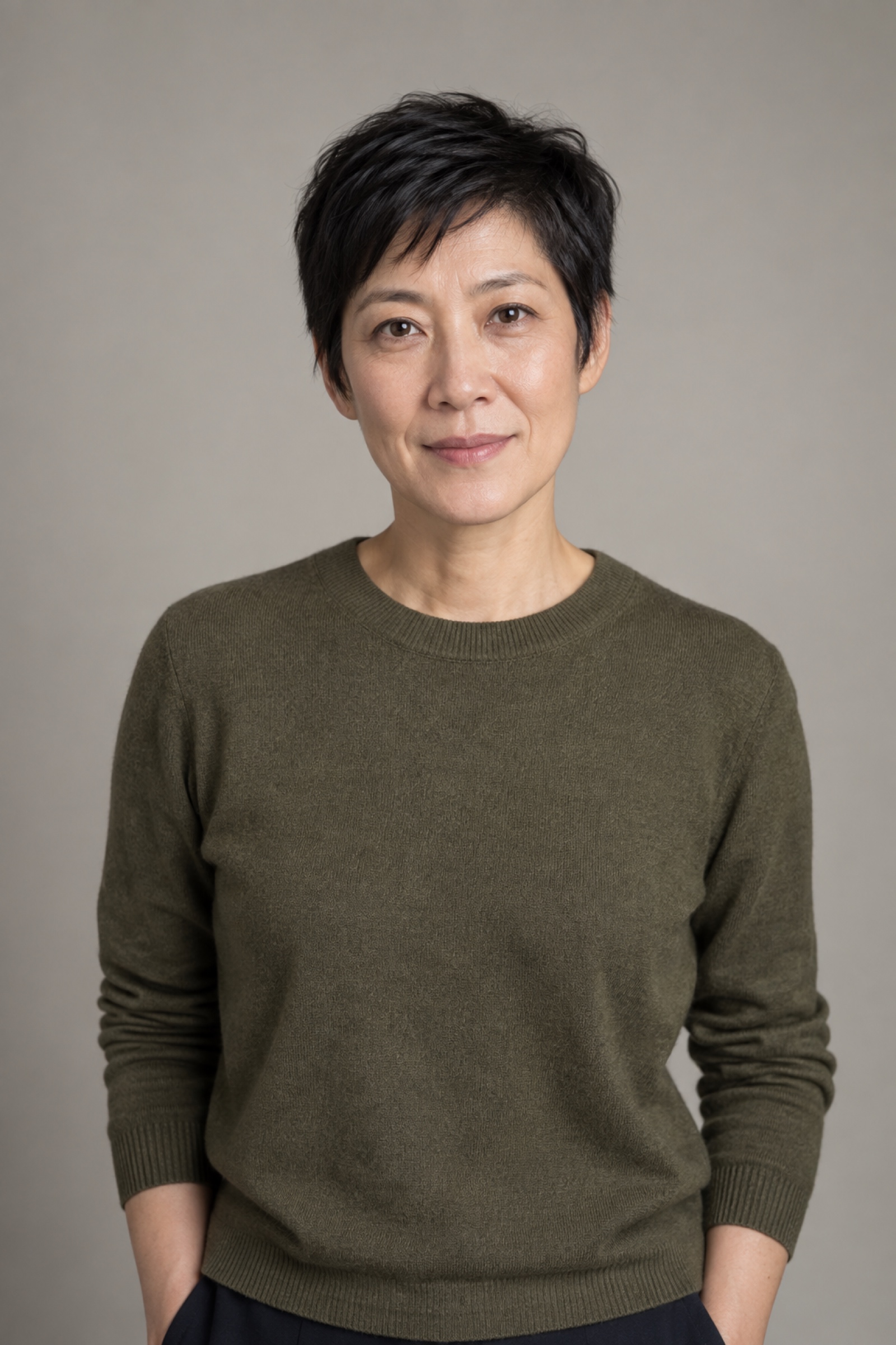Studio portrait of a woman in their 40s with short dark pixie haircut, wearing an olive sweater, against a warm grey background.