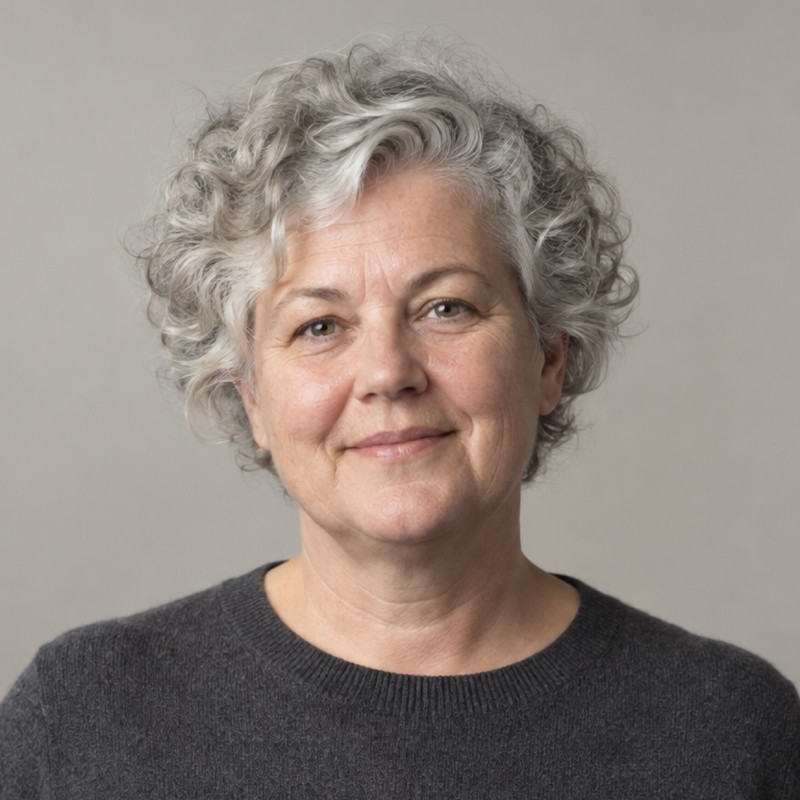 Head and shoulders of a woman in their 50s with short naturally silver curly hair, wearing a charcoal sweater, against a warm grey background.