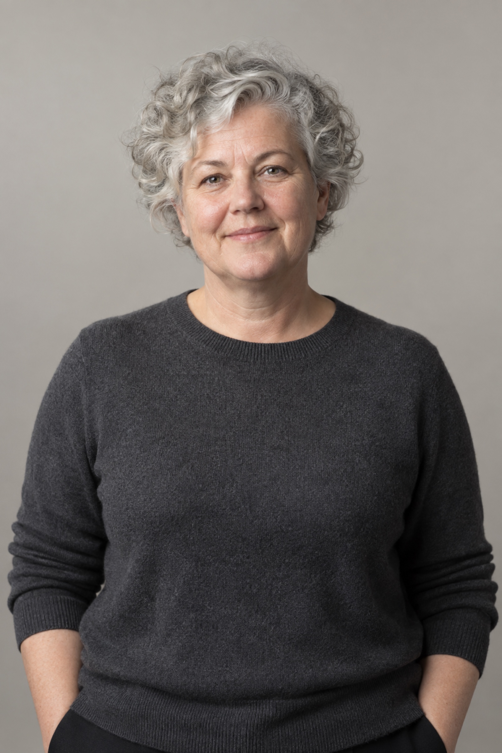 Studio portrait of a woman in their 50s with short naturally silver curly hair, wearing a charcoal sweater, against a warm grey background.