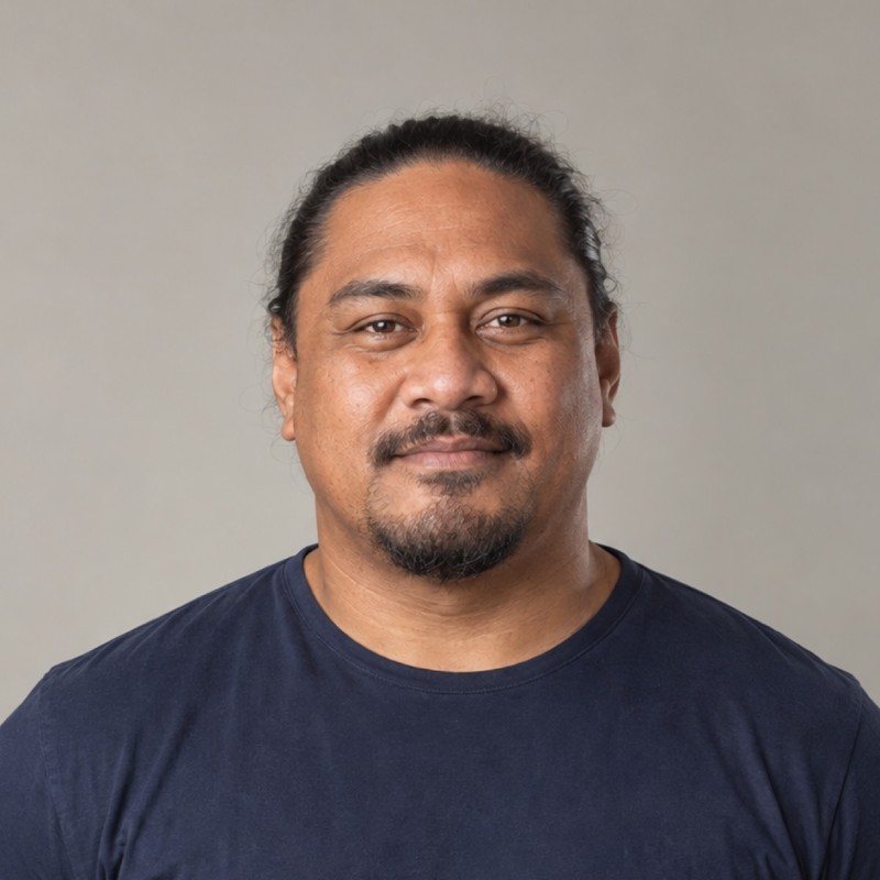 Head and shoulders of a man in their 40s with long dark hair tied back, wearing a navy shirt, against a warm grey background.