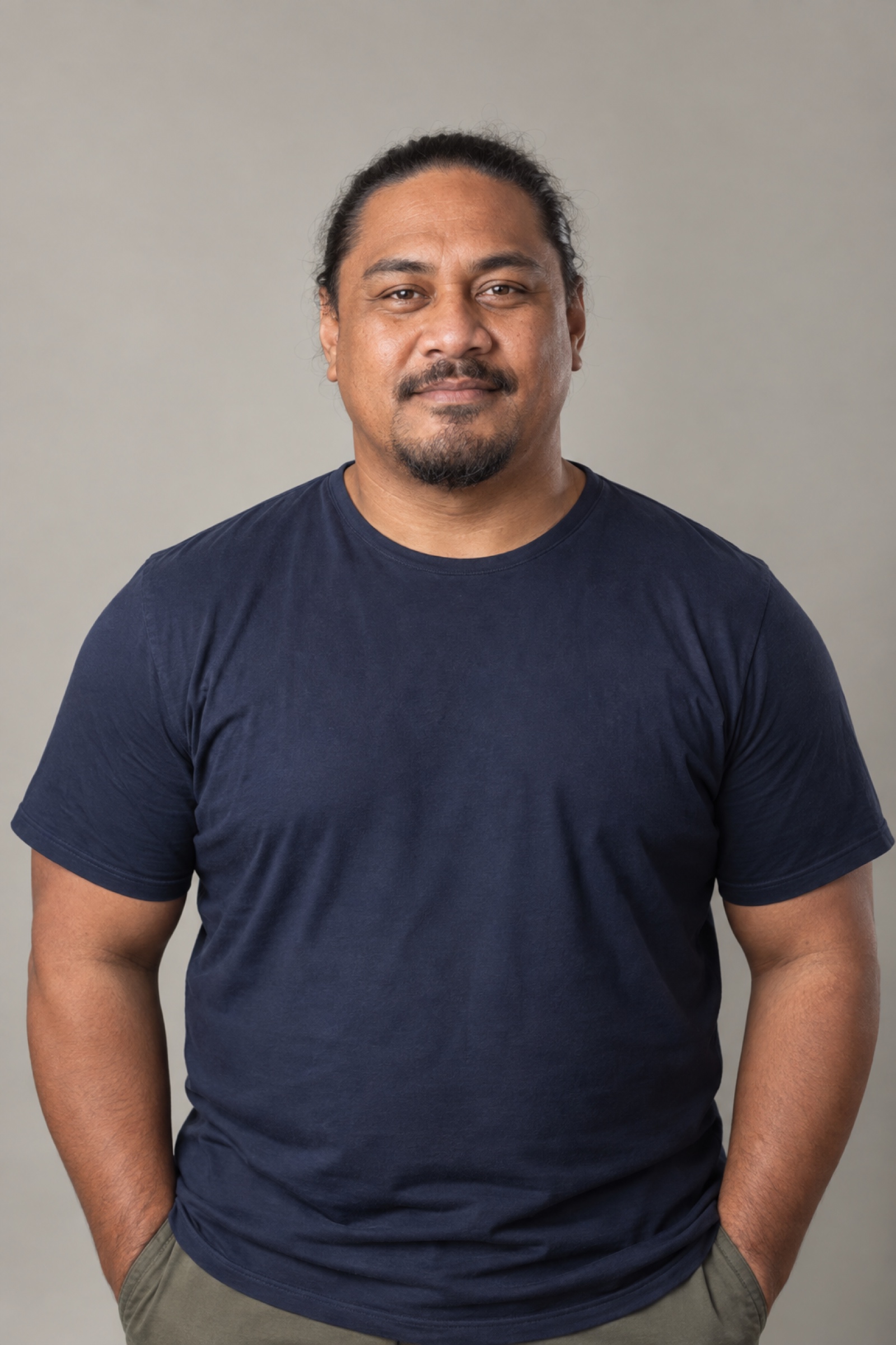 Studio portrait of a man in their 40s with long dark hair tied back, wearing a navy shirt, against a warm grey background.