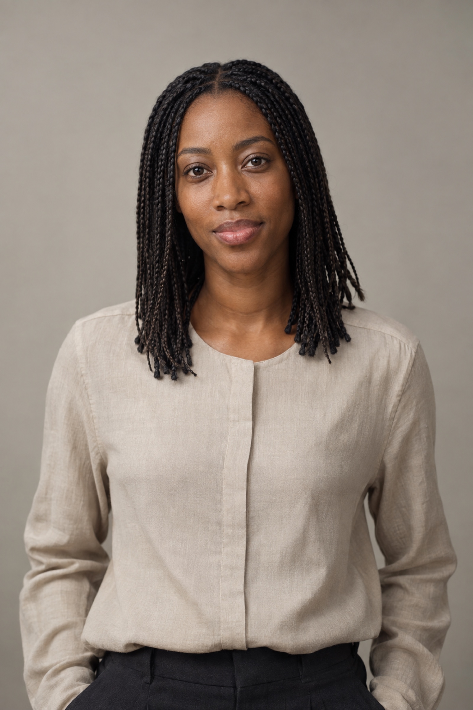 Studio portrait of a woman in their 30s with shoulder-length neat microbraids, wearing a stone blouse, against a warm grey background.