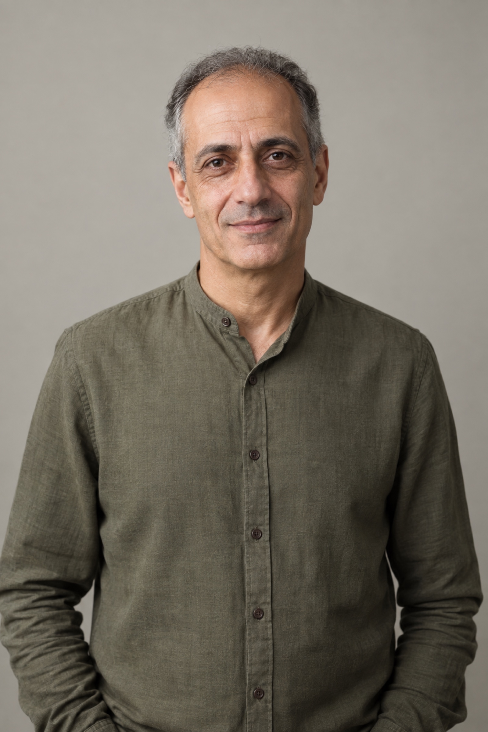 Studio portrait of a man in their 50s with receding short salt-and-pepper hair, wearing an olive shirt, against a warm grey background.
