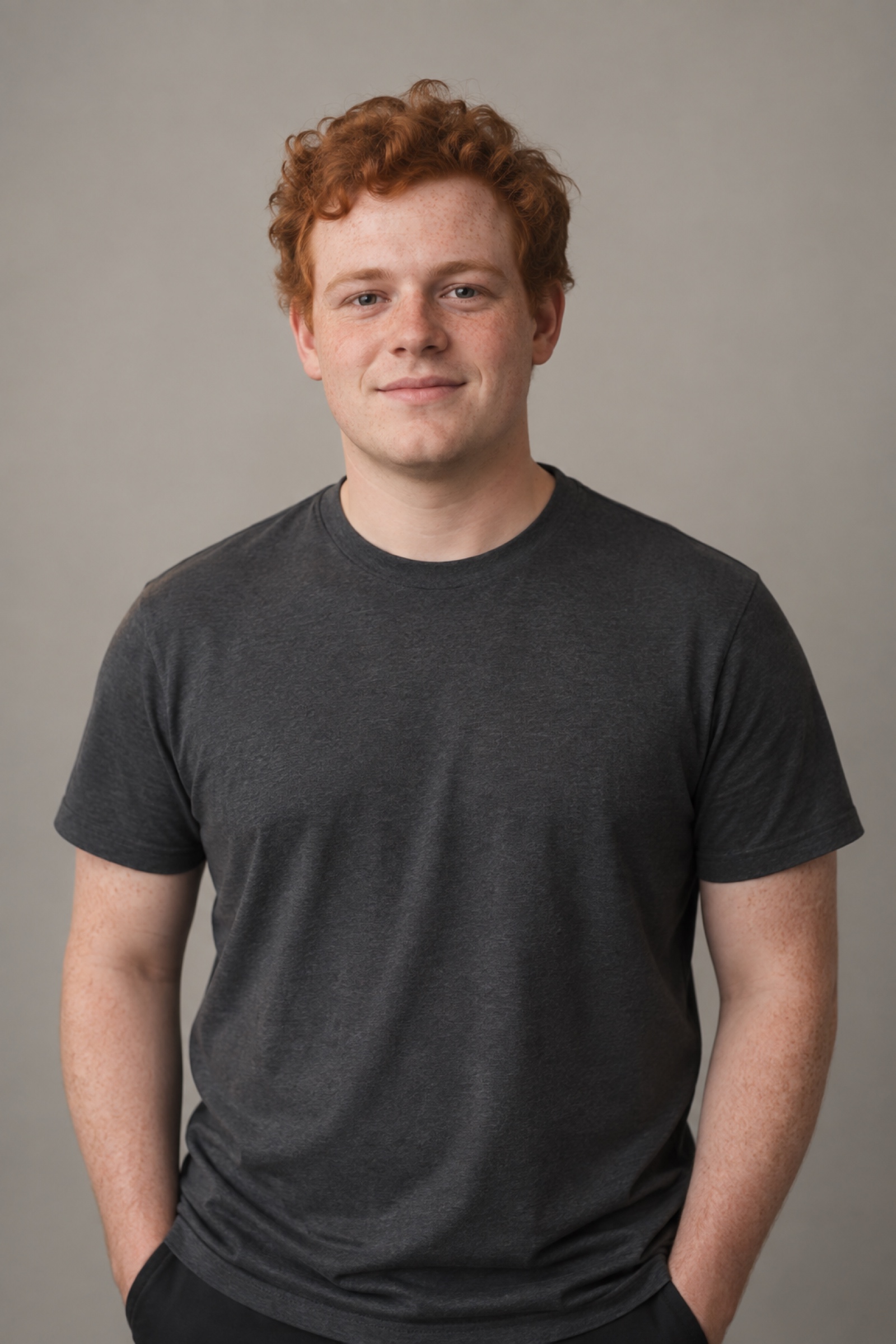 Studio portrait of a man in their 20s with short ginger curls, wearing a charcoal shirt, against a warm grey background.