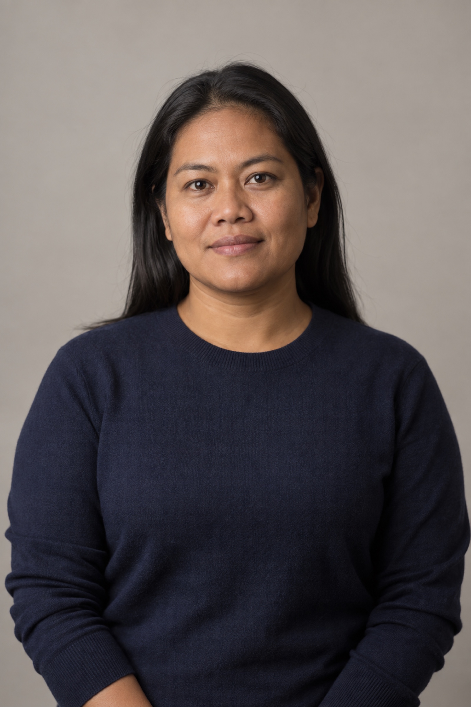 Studio portrait of a woman in their 40s with long straight dark hair resting behind the shoulders, wearing a navy sweater, against a warm grey background.
