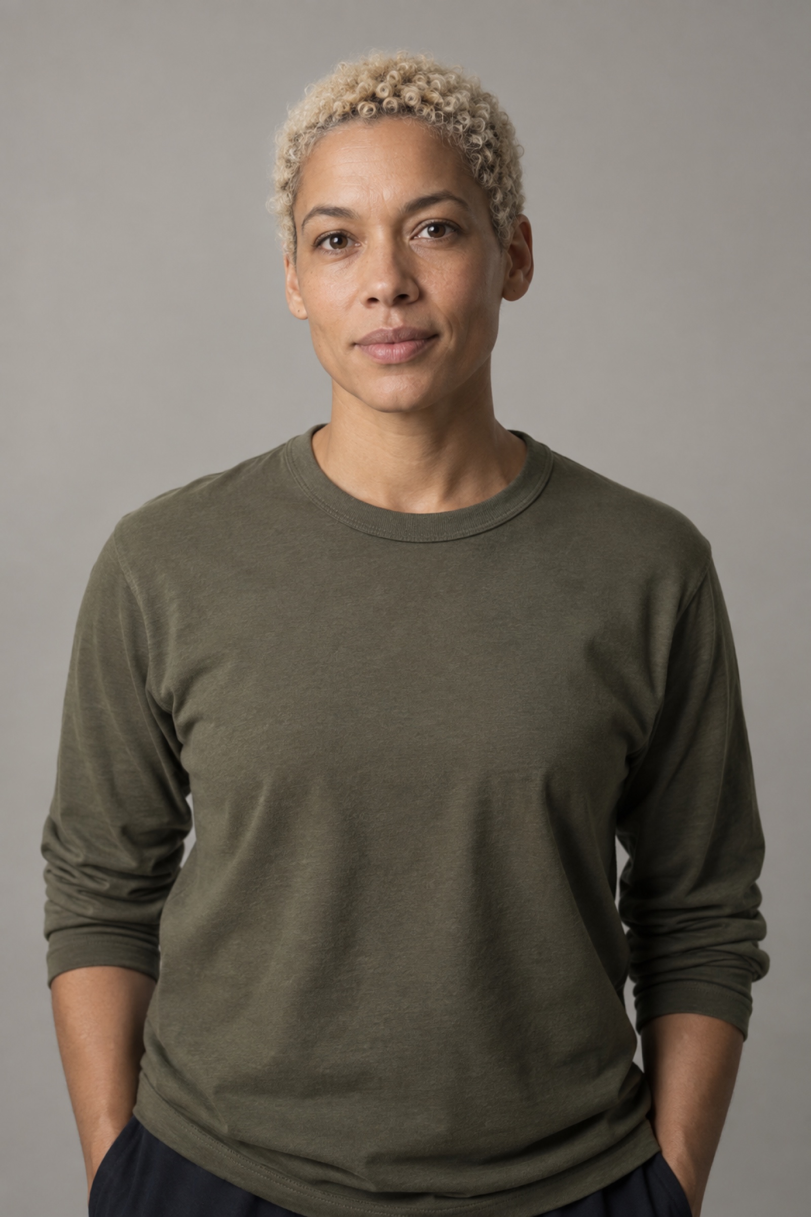 Studio portrait of a person in their 30s with close-cropped bleached-blond natural curls, wearing an olive shirt, against a warm grey background.